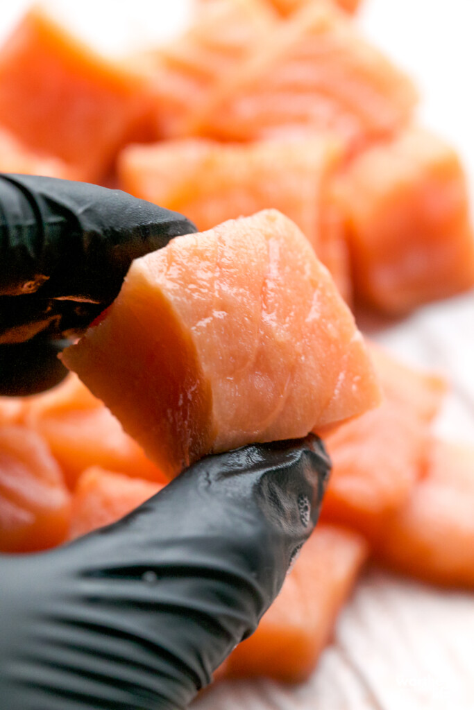 man with a black food glove on holding a fresh piece of cubed salmon