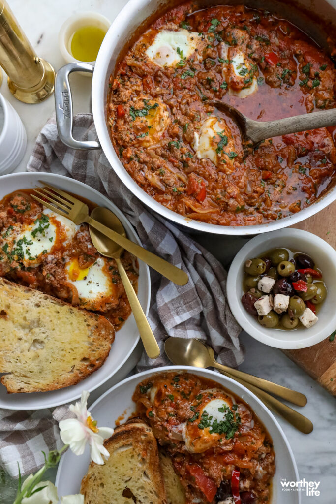 Lamb Shakshuka in a pan, as well as toasted sourdough bread with a bowl of olives